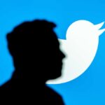 Mudanças do Twitter afastam usuários e anunciantes. A imagem mostra a sombra de Elon Musk, CEO do Twitter, à frente do logo da rede social: um pássaro branco sobre um fundo azul claro.